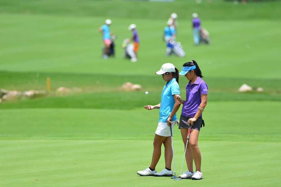 果岭边的博弈与情谊青少年高尔夫赛事中的专注时刻与团队精神 两位女球员在果岭上安静等候,沐浴在阳光下,展现出耐心与优雅并存的赛场风度。