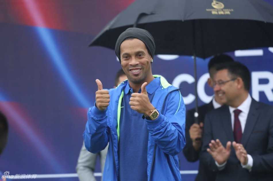 罗纳尔迪尼奥被一群小球员环绕，他开怀大笑，双手高举比出点赞手势，画面充满欢乐与活力。