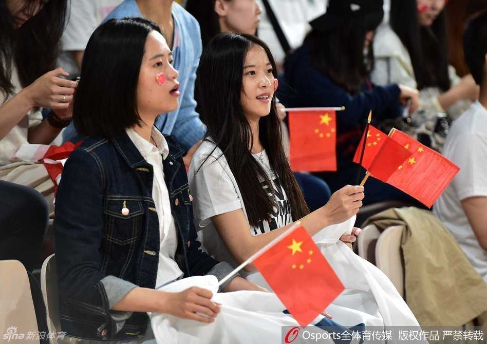 镜头聚焦于一位正在整理妆容的中国美女球迷，她修长的手指轻轻撩起乌黑的长发，眼神专注而温柔，随后将一枚五星红旗贴纸端正地贴在脸颊上，青春洋溢的脸上写满了对女篮的期盼与祝福。