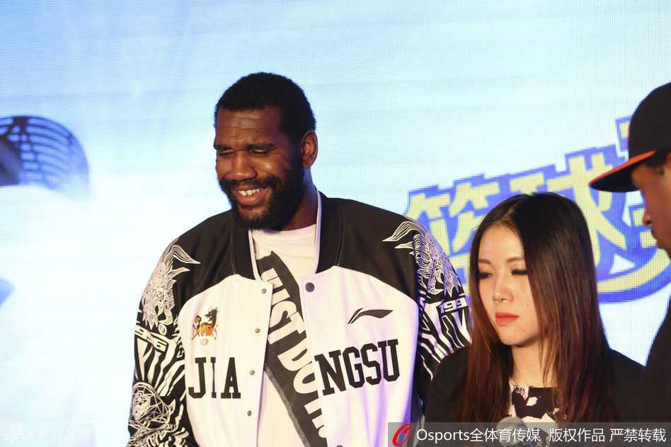 奥登双手抱胸站立台上，脸上绽放出灿烂笑容，身旁的胡雪峰正在与工作人员交谈，奥登被现场气氛感染，眼中闪烁着轻松愉悦的光芒。