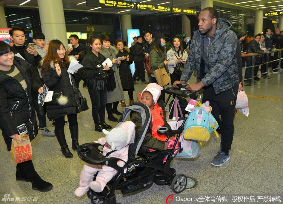 哈德森弯着腰，双手稳稳地推着婴儿车，妻子穿着白色羽绒服跟在身侧，一家三口在人群中缓慢穿行，画面温馨有爱。