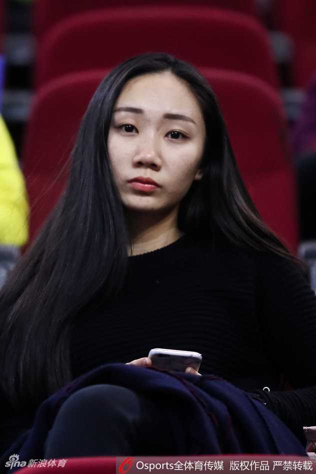 看台上的她，身着简洁的白色上衣，手持一杯饮料，正侧身与同伴交流。身后的赛场灯光映照在她精致的侧脸上，让这一刻的观赛时光显得既惬意又充满活力。