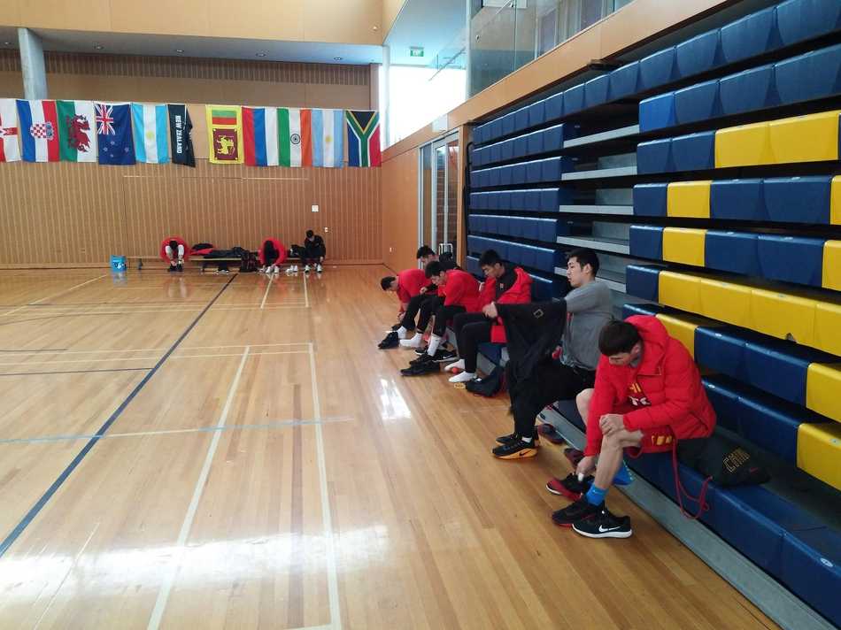 场馆外，阴雨绵绵，一位队员透过窗户凝望着远方的雨景，棉服的帽子还戴在头上，仿佛在静静思索着下一场比赛的战术。