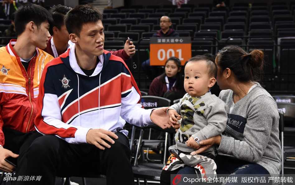 周鹏与刘美各抱着一个孩子，孩子们的目光被场内激烈的比赛所吸引，周鹏则微微侧身与妻子交流，仿佛在分享着对某位年轻球员表现的看法