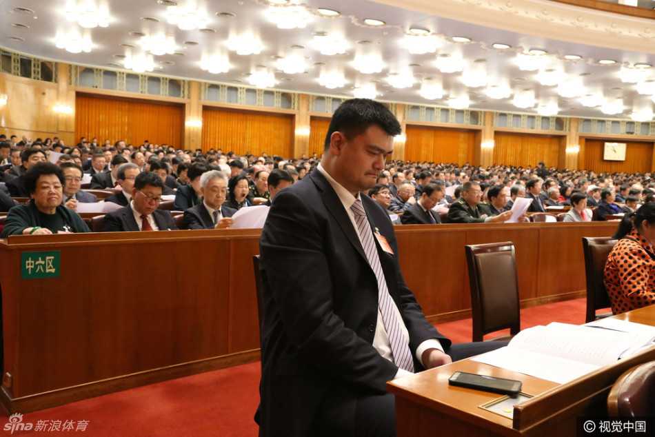 在人民大会堂内的休息区，姚明微微低头认真阅读手里的会议文件，巨大的身高差让他身边的普通工作人员显得十分娇小，画面充满反差萌。