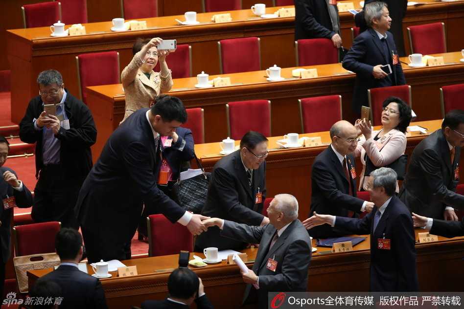 姚明结束发言后回到座位，他面带微笑与旁边的委员进行交流，神态放松，远处会场的灯光璀璨，背景中能看到其他委员专注的神情，这一刻充满了履职后的释然与继续前行的力量。