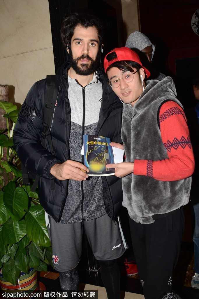 一位戴着眼镜的年轻女球迷终于如愿以偿，她激动地站在一位高大的同曦队员身旁，双手比出胜利的‘V’字，脸上的笑容比身后的圣诞装饰更加灿烂，队员也微微俯身配合，画面定格在温馨的瞬间。