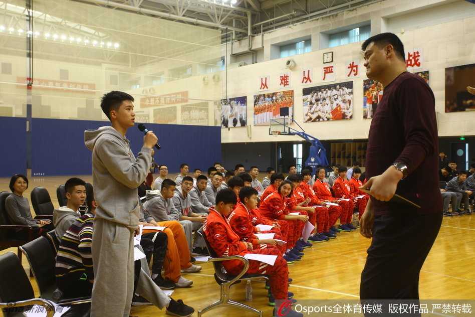 姚明坐在场边小板凳上侃侃而谈，周围坐满了身穿国家队队服的青少年球员，他眼神中充满鼓励与期待，仿佛要将自己所有的经验都倾囊相授给下一代。