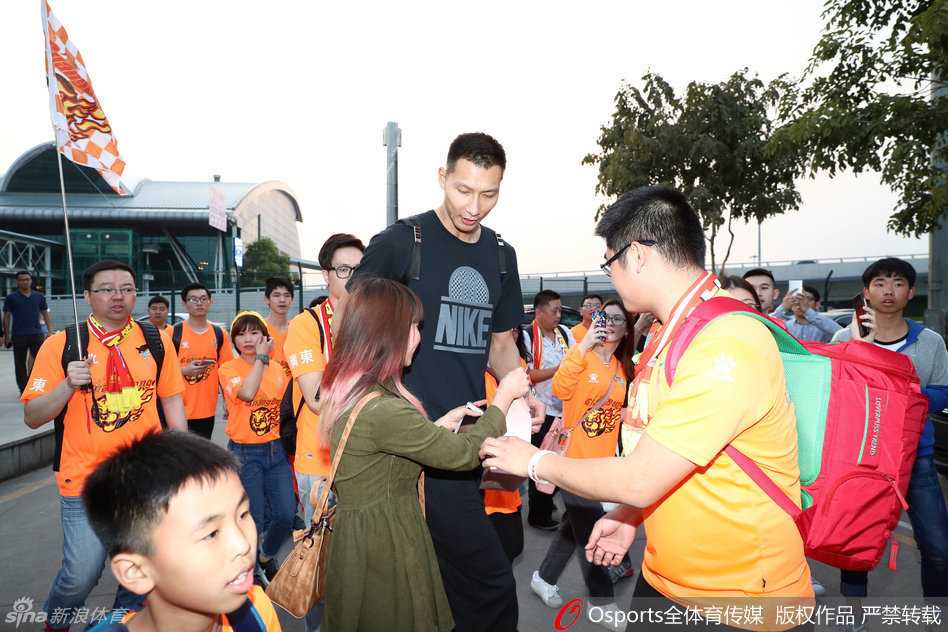 身穿红色外套的易建联神情专注，正低头为热情的球迷递上的球衣签名，眼神中带着一丝疲惫却满是耐心。