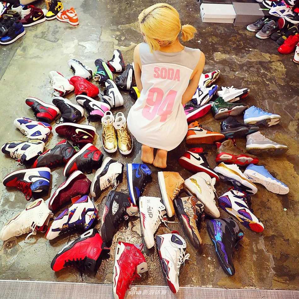 在暖色调的灯光下，DJ Soda坐在堆积如山的球鞋中间，她身穿黑色T恤和破洞牛仔裤，手里拿着一双球鞋仔细端详，身旁色彩斑斓的鞋盒仿佛是她最珍贵的宝藏库。