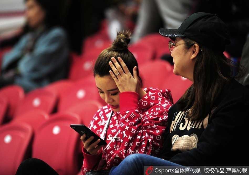 李根妻子王星洋扎着清爽丸子头，侧颜轮廓精致，身穿休闲外套坐在看台上，目光越过人群投向赛场方向，眼神中充满期待。