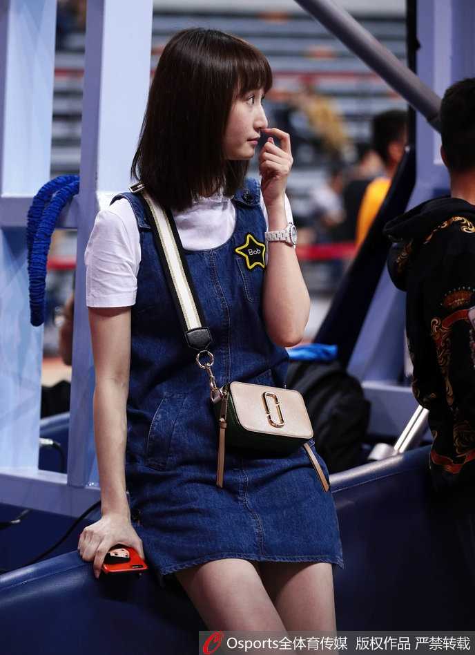 篮球场边的最美风景:周鹏妻子刘美见证“粤川战”的柔情时刻 刘美在赛场边低头查看手机,嘴角带着一抹温柔的笑意,牛仔裙的简约设计衬托出她清新脱俗的气质,与身后紧张的比赛氛围形成有趣对比。