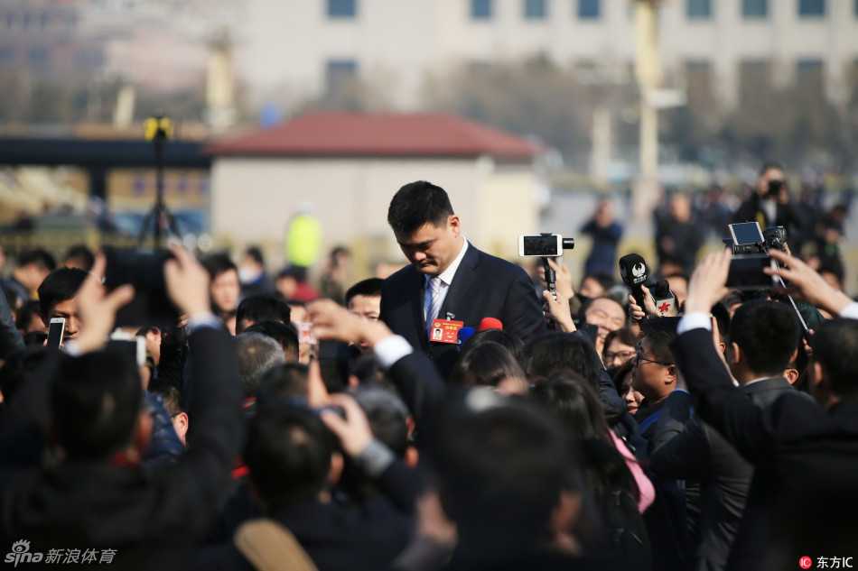 姚明弯腰俯身，将耳朵凑近一位拿着话筒的小记者，耐心倾听提问，巨大的身高差构成了一幅温馨有爱的画面