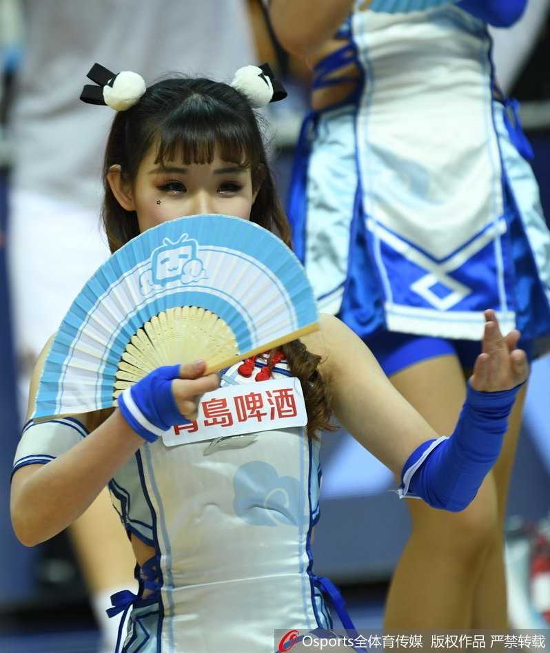 一位啦啦队女孩的特写，她微微侧身，对着镜头露出治愈系的甜笑，手上拿着一个印有可爱卡通形象的应援手幅，背景是虚化的球场和观众，整个画面充满了青春与活力的气息。