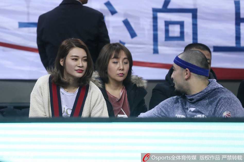 李根妻子与西热力江在场边低声交谈，两人神情专注，西热力江比划着手势，似乎正在分享着关于场上局势的什么秘密。