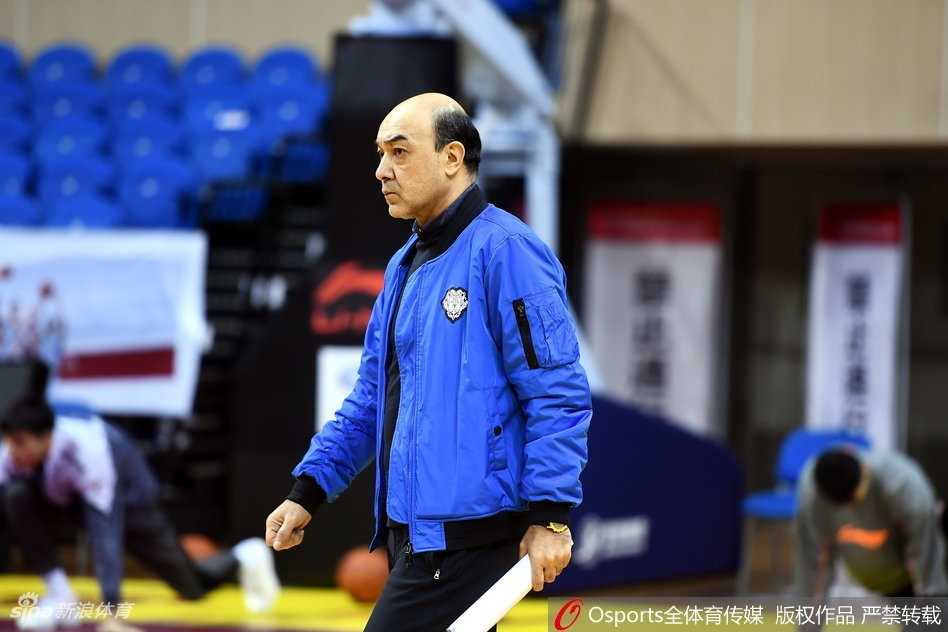 训练间隙，阿的江指导双手叉腰，神情略显疲惫却依然坚毅，他的背后是刻苦训练的年轻队员，一老一少构成了一幅传承的画面。