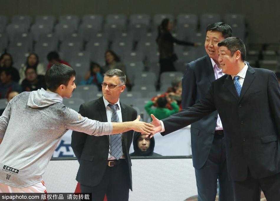 王治郅双手背后，神情专注地注视着场上的八一队年轻队员，身后是正在热身的对手，他的身影显得沉稳而坚定，宛如定海神针。