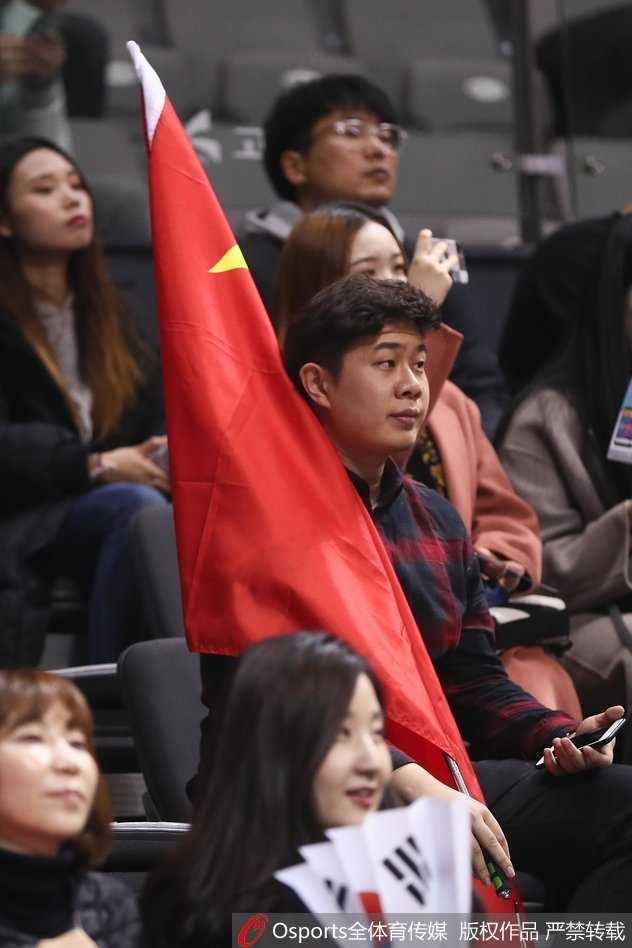 几位中国球迷并肩站立，其中一人高举着五星红旗，他们的目光紧紧锁定在赛场中央。周围的韩国观众神情各异，红色的旗帜在这一片陌生的色彩中显得醒目而坚定。