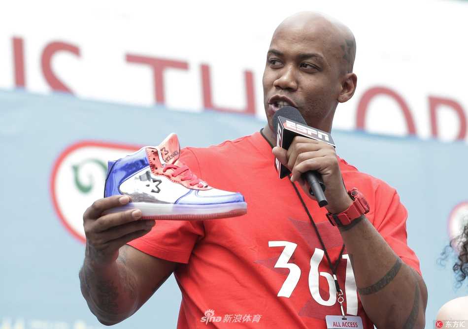 马布里现身纽约科尼岛热狗大赛现场，身穿深色紧身运动衣搭配个人品牌球裤，面带微笑望向镜头，背景是熙熙攘攘的围观人群和美国国旗装饰。